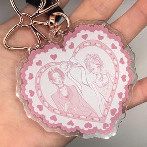 May include: A clear acrylic keychain with a pink heart-shaped design. The heart features a couple illustration and is surrounded by a scalloped edge and small hearts. The keychain has a rose gold bow-shaped clasp.