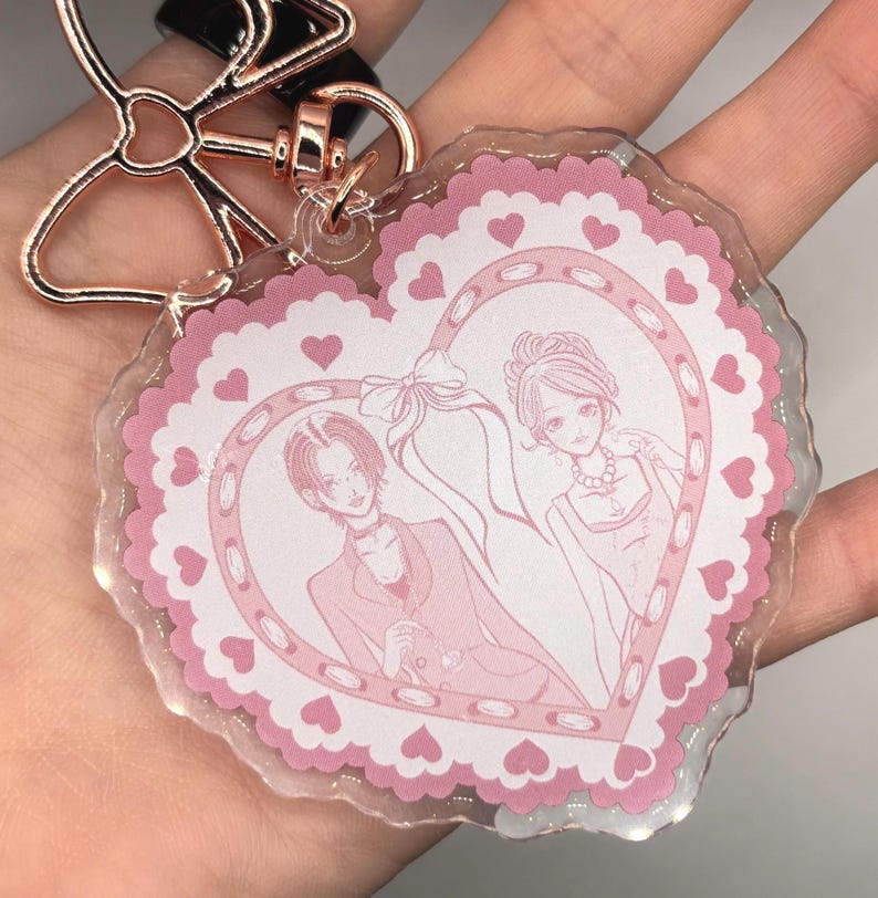 May include: A clear acrylic keychain with a pink heart-shaped design. The heart features a couple illustration and is surrounded by a scalloped edge and small hearts. The keychain has a rose gold bow-shaped clasp.