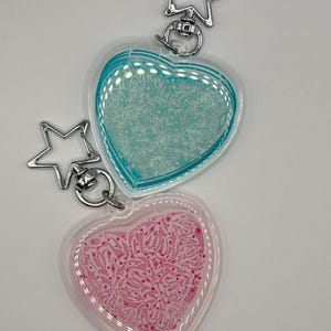 May include: Two heart-shaped keychains with silver star-shaped clasps. One keychain has a teal interior design, and the other has a pink interior design. Both keychains have a clear border.