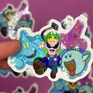 May include: A holographic sticker featuring Luigi from the Super Mario Bros. series, surrounded by ghosts. Luigi is depicted with a vacuum, and the sticker has a shiny, iridescent finish. The sticker is held against a pink background.