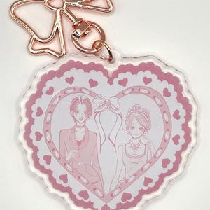 May include: A heart-shaped keychain with a pink and white design. The keychain features a pink border with heart details and a central illustration of two figures. The keychain has a rose gold bow-shaped clasp.
