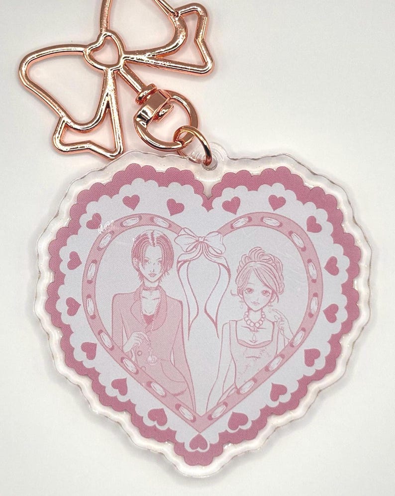May include: A heart-shaped keychain with a pink and white design. The keychain features a pink border with heart details and a central illustration of two figures. The keychain has a rose gold bow-shaped clasp.