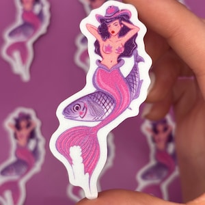 May include: A sticker featuring a mermaid with a fish tail and a cowboy hat. The mermaid has dark hair, pink lips, and is wearing a pink top. The fish tail is pink and purple. Other mermaid stickers are in the background.