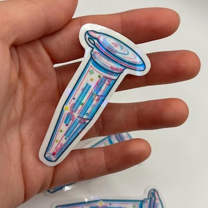 May include: A holographic sticker featuring a stylized, colorful laboratory vial design. The sticker has a white border and is decorated with blue, pink, and yellow accents. The vial shape is elongated with a rounded top.