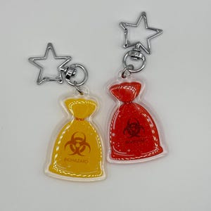 May include: Two keychains with a bag design. One is yellow with a biohazard symbol and the word "BIOHAZARD". The other is red with a biohazard symbol. Both have a silver star-shaped clasp.