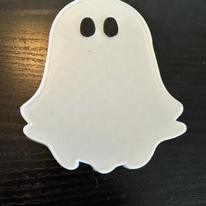 May include: A white ghost-shaped decoration with two black oval eyes. The ghost has a smooth, rounded shape with a slightly wavy edge. This Halloween-themed decoration is perfect for adding a spooky touch to any space.