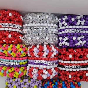 Könnte beinhalten: Eine Sammlung bunter Perlenarmbänder. Die Armbänder sind in verschiedenen Farben erhältlich, darunter Rot, Weiß, Lila, Gelb und Blau. Jedes Armband hat einen Namen, der in weißen Buchstabenperlen geschrieben ist. Die Armbänder sind gestapelt.