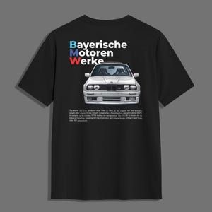 Puede incluir: Camiseta negra con una ilustración blanca de un BMW M3 clásico. La camiseta tiene el texto "Bayerische Motoren Werke" en azul, blanco y rojo. Debajo del coche hay texto sobre la historia del coche.