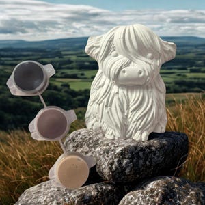May include: A white Highland cow sculpture with textured fur sits on a stone. Three small paint pots in grey, brown, and beige are connected and to the left of the cow. The background shows a scenic landscape.