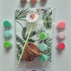 May include: A paint-at-home kit featuring a small terracotta pot, a paintbrush, and a fabric sheet with a green leaf pattern. The kit includes various paint colours in small containers, including brown, orange, green, pink, and purple.