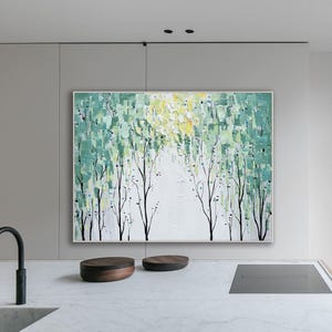 May include: An abstract landscape painting with a white frame. The artwork features a forest scene with green and yellow brushstrokes, and black tree trunks. The painting is displayed above a white countertop in a modern kitchen setting.