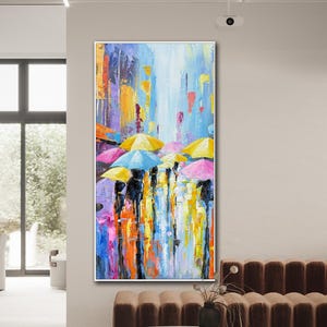 May include: An abstract painting depicts a rainy street scene with figures holding umbrellas. The artwork features a vibrant palette of blues, yellows, pinks, and oranges, creating a colorful and dynamic composition. The painting is framed in white.