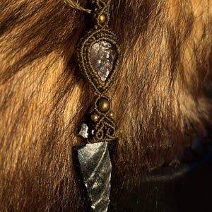 May include: A handcrafted macrame necklace featuring a dark gray arrowhead pendant and a light purple crystal. The necklace has a bronze-colored cord and accents. The pendant is approximately 2 inches long.