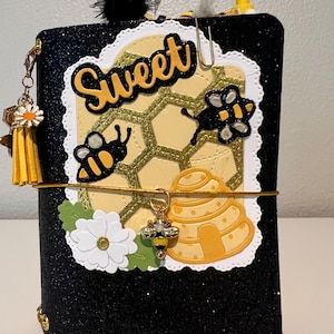 May include: A black glittery notebook cover featuring a central die-cut panel with a yellow honeycomb pattern, the word Sweet in black script, two bee cutouts, a yellow beehive, a white flower, and a gold elastic band with a bee charm.