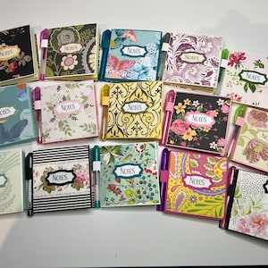 May include: A collection of fourteen square notebooks with various floral, geometric, and butterfly patterns on the covers. Each notebook features a white label with the word Notes printed in black and includes a matching pen held in a side loop.