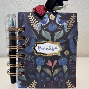 May include: A spiral-bound notebook with a dark floral pattern featuring blue birds, green leaves, and roses. A gold-trimmed label in the center reads "Beautiful". A black pen is held by the gold wire binding, and a black and red ribbon sits on top.