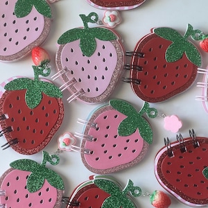 May include: A collection of strawberry-shaped notebooks in various shades of pink and red. Each notebook features a glittery green stem and a spiral binding. Some notebooks have small charms attached, including a strawberry and a flower.