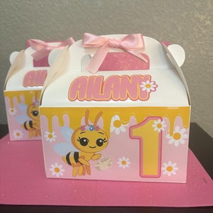 May include: Two white party favor boxes with pink accents. Each box features a cartoon bee, the number 1, and the name "AILAN" in pink. The boxes are decorated with daisies and topped with pink bows.