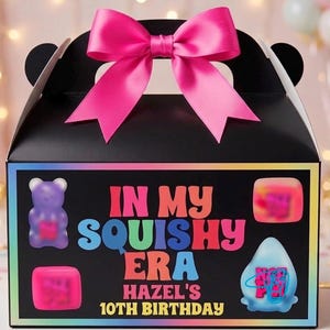 May include: A black gable box with a large pink satin bow on top. The front features the text "IN MY SQUISHY ERA HAZEL'S 10TH BIRTHDAY" in colorful letters, surrounded by illustrations of a purple bear, a pink square, and a blue triangular shape.