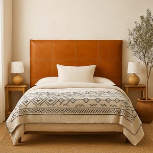 May include: A bed with a tan leather headboard, a cream-colored pillow, and a patterned blanket. Two small wooden side tables with lamps flank the bed. A potted plant is on the right side of the bed.