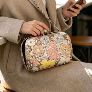 Op de afbeelding: Een rechthoekige clutch met een dicht, sprankelend oppervlak van lovertjes met een bloemenpatroon in roze, gele en witte tinten. De tas is voorzien van een metalen schouderketting en een decoratieve sluiting bezet met kristallen.