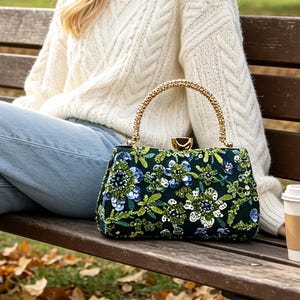 Könnte beinhalten: Eine dunkelgrüne, strukturierte Handtasche mit einem komplexen Blumenmuster aus blauen, grünen und weißen Pailletten und Perlen. Die Tasche hat einen goldfarbenen Metallrahmen, einen goldfarbenen Schnappverschluss und einen strukturierten Perlenhenkel.