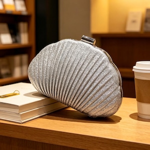 May include: A silver glittery clutch bag shaped like a seashell with a metallic clasp closure. The bag features a textured, ribbed surface that mimics the appearance of a shell, providing a shimmering finish to the overall design of the accessory.