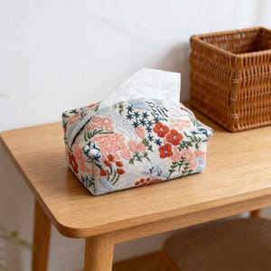 Op de afbeelding: Geborduurde tissuebox met een bloemenpatroon in rood, roze, blauw en groen. De hoes staat op een houten tafel, met een rieten mand op de achtergrond. De tissuebox is vierkant.