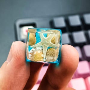 May include: A clear, cube-shaped resin keycap with a starfish, seashells, and sand suspended in blue resin. The keycap is held in a person's hand, with a keyboard in the background. The keycap is a unique decorative item.