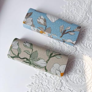 以下が含まれることがあります： 花柄の刺繍が施された2つの長方形のケース。1つは水色の背景に白と茶色の花模様が描かれています。もう1つはベージュの背景に白と緑の花のデザインが施され、黄色のアクセントが加えられています。
