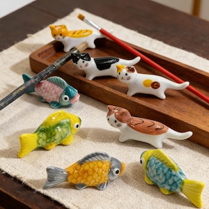 Op de afbeelding: Een verzameling keramische eetstokjessteunen in de vorm van katten en vissen in verschillende kleuren, waaronder oranje, zwart, wit, geel en blauw. De eetstokjessteunen zijn gerangschikt op een houten dienblad met penselen.