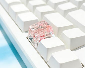 May include: A clear resin mechanical keyboard keycap featuring embedded small pink floral elements, positioned among standard solid white keycaps on a white keyboard base. The keycap has a smooth, glossy finish and a square shape.