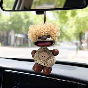 May include: A whimsical car charm in the shape of a cartoon character. The charm features a light brown, textured body, dark brown arms and legs, and a curly, light brown head of hair. The mouth is open and red. The charm hangs from a brown string.