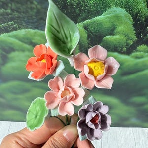 May include: A cluster of ceramic flowers in shades of orange, pink, and purple, with green leaves. The flowers have yellow centers and are held in a hand. The background is a blurred green landscape.
