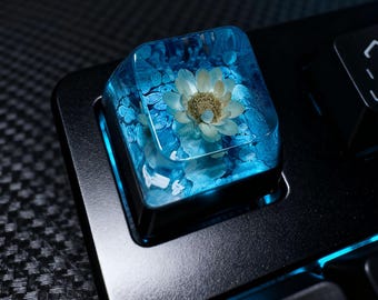 May include: A custom keyboard keycap with a clear resin body, encasing a dried white flower and blue decorative elements. The keycap is set on a black keyboard, with a focus on the unique, artistic design. The keycap is a square shape.