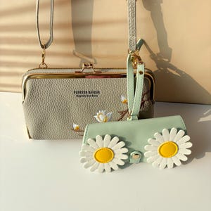 May include: A light grey crossbody bag and a matching mint green wallet. The crossbody bag has a kiss-lock closure and the text "FOREVER SAVOUR". The wallet is decorated with two large daisy appliques.