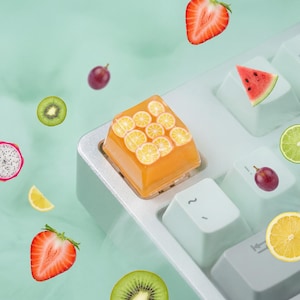 Könnte beinhalten: Eine durchscheinende orangefarbene Harz-Tastenkappe mit eingebetteten Miniatur-Zitrusfrüchtescheiben auf einer hellen mintgrünen mechanischen Tastatur, umgeben von schwebenden Abbildungen von Erdbeeren, Kiwi, Wassermelone, Weintrauben und Zitronenscheiben.