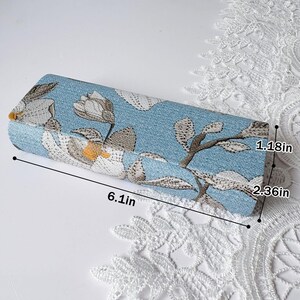 以下が含まれることがあります： 青い布製の長方形ケースで、黄色い中心を持つ白い花の刺繍が施されています。画像内の測定ラベルが示す通り、ケースの長さは15.49cm、幅は5.99cm、高さは3cmです。