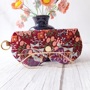 May include: A burgundy embroidered glasses case with a gold clasp and button closure. The case features a floral design in orange, pink, and white. Gold-rimmed glasses rest in front of the case. A vase of flowers is in the background.