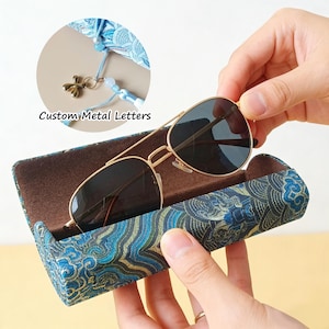 Puede incluir: Gafas de sol aviador con montura dorada y lentes oscuras en un estuche estampado. El estuche es azul y verde con un diseño de olas. El texto "Custom Metal Letters" es visible.