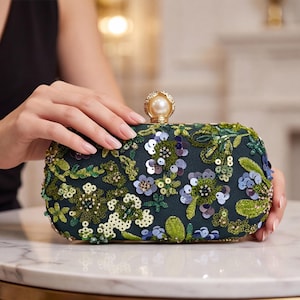 May include: A dark navy blue rectangular clutch bag featuring intricate floral embroidery with green and blue sequins, beads, and threadwork. The bag is topped with a gold-toned metal clasp featuring a large, round, cream-colored faux pearl.
