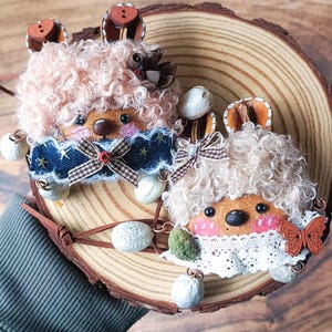 May include: Two handcrafted hedgehog decorations with light brown, curly fur and decorative features. One has a denim-style bib with star details and a bow. The other has a white lace bib and a butterfly. Both have button ears and are on a wooden slice.