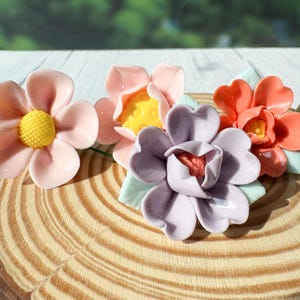 Peut inclure: Quatre fleurs en céramique aux tons rose, pêche et violet, avec des centres jaunes et rouges. Les fleurs sont disposées sur une tranche de bois avec des motifs de grain visibles. Les fleurs ont des feuilles vertes.