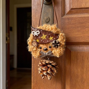May include: A whimsical, handmade door hanging featuring a plush, curly-haired bear with a brown leather hat adorned with gold stars. A decorative bow and a pinecone complete the design. The hanging is suspended from a brown cord.