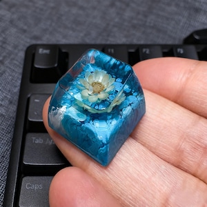 Könnte beinhalten: Eine quadratische Tastenkappe mit einer Blume, die in klarem Harz eingeschlossen ist. Das Harz ist ein leuchtendes Blau mit weißen Wirbeln, was ein einzigartiges, künstlerisches Design ergibt. Die Tastenkappe befindet sich auf einer schwarzen Tastatur.