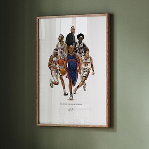 May include: A framed art print featuring a group illustration of the 2003-2004 Detroit Pistons basketball team in action. The players wear white and blue uniforms, with one player in the center wearing blue. Text at the bottom reads 2003-2004 NBA CHAMPIONS // DETROIT PISTONS.