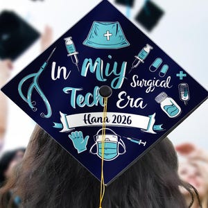 May include: A graduation cap with a navy blue background and medical-themed illustrations. The cap reads "In Miy Surgical Tech Era Hana 2026" in white and teal lettering, with a yellow tassel hanging down. The image is taken from behind a graduate.