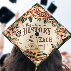 May include: A graduation cap with a floral design and the text "Born to make HISTORY...and TEACH". The cap features butterflies, roses, and a stack of books. A yellow tassel hangs from the cap. The name "Ms. Selena 2026" is also on the cap.