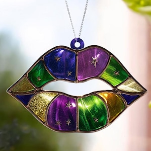 May include: A stained glass lip-shaped suncatcher with a silver chain. The lips are segmented into colorful sections of purple, green, gold, and blue, each with star accents. The suncatcher hangs in front of a window.
