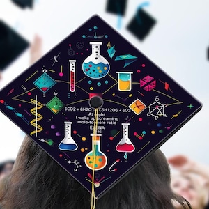 May include: A graduation cap with a dark blue background, decorated with colorful science-themed illustrations of beakers, test tubes, and chemical formulas. The text reads "6CO2 + 6H2O C6H12O6 + 6O2 At night I woke up screaming mole-to-mole ratio ELENA 26". A gold tassel hangs from the cap.